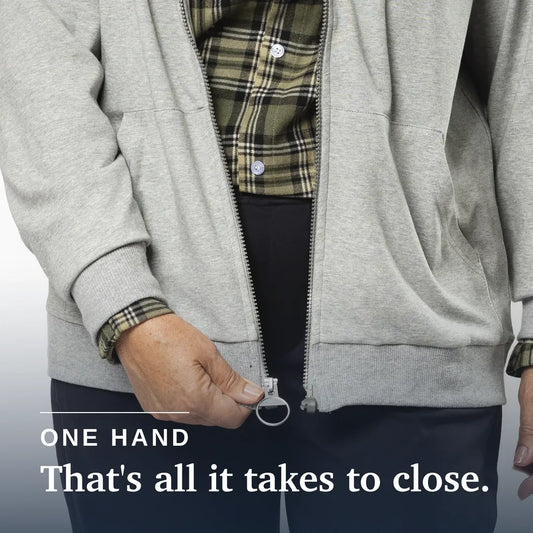 An older woman stands against a plain white studio background wearing a light gray zip-up hoodie over a green plaid shirt, dark navy pants, and cream-colored sneakers. #Grey