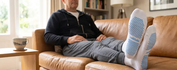 A man reclining on a brown leather couch with his legs stretched out, wearing white socks with blue textured grip soles. The setting is a cozy living room with a wooden side table, a mug, and a bookshelf in the background.