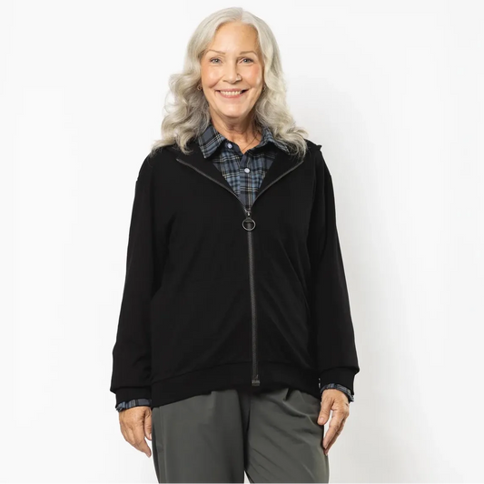 An older woman with gray hair stands against a white studio background wearing a black zip-up hoodie over a blue plaid shirt, dark gray pants, and beige shoes. #Black