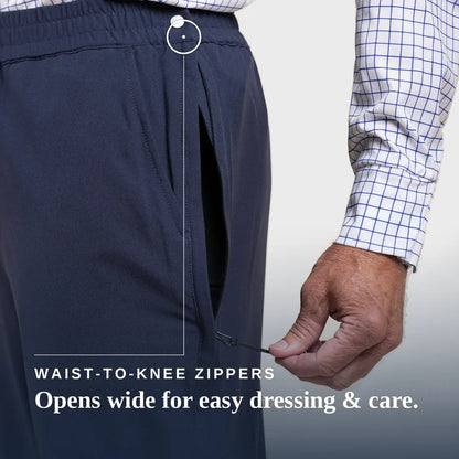 Close-up of a person wearing dark navy pants with a hand demonstrating the wide opening of the waist-to-knee zipper. A white and blue checkered shirt sleeve is visible, and text reads 'WAIST-TO-KNEE ZIPPERS' and 'Opens wide for easy dressing & care. Joe & Bella CareZips® Men's Access Pants #Dark Navy