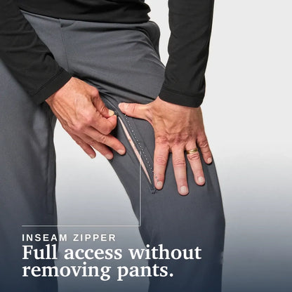 Close-up of a person wearing dark gray pants and a black long-sleeve top, demonstrating an inseam zipper that allows full access without removing the pants. Text at the bottom reads 'INSEAM ZIPPER Full access without removing pants. Joe & Bella CareZips® Men's Access Pants #Slate Grey