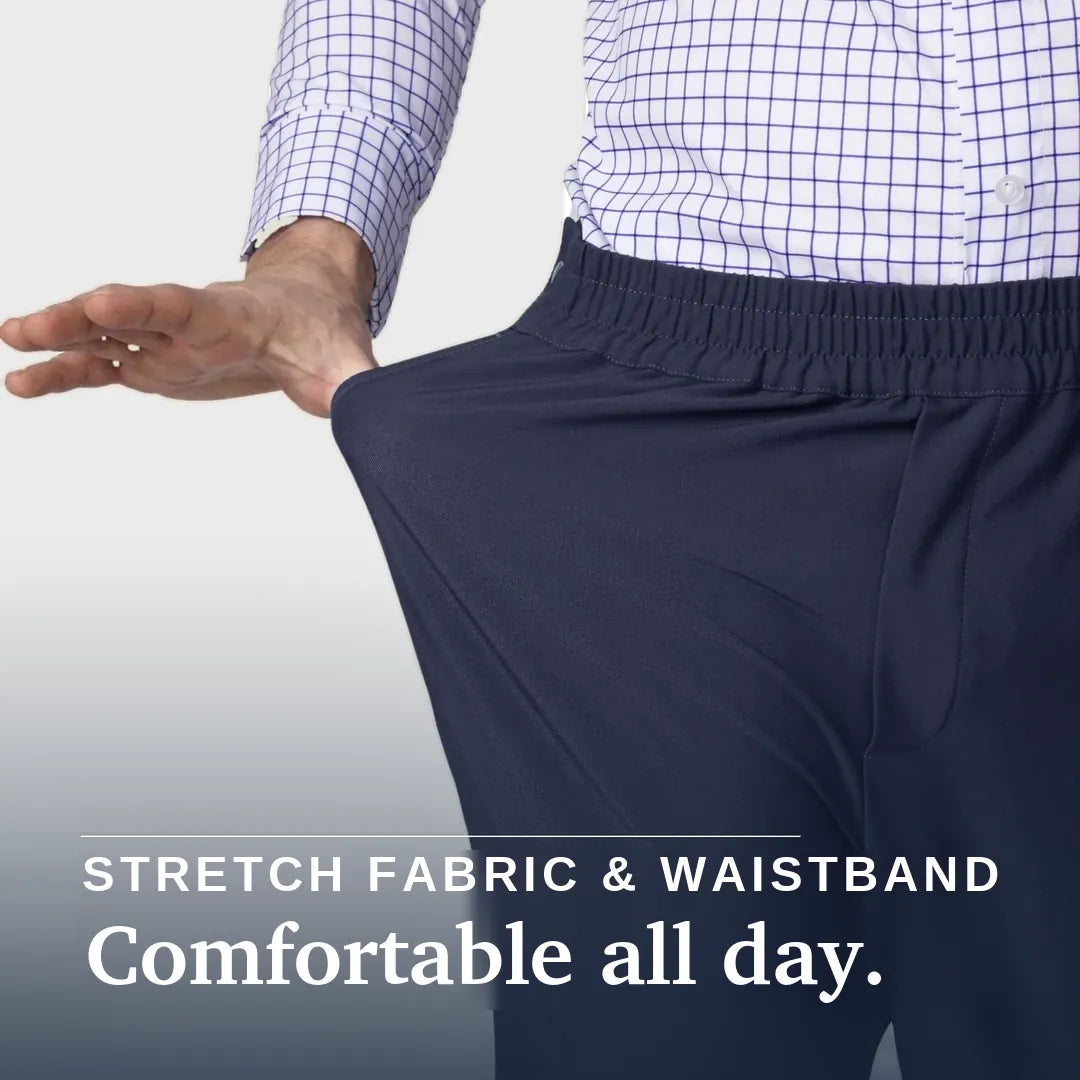 A close-up of a person wearing dark navy elastic-waist pants and a white button-up shirt with a blue grid pattern, stretching the fabric of the pants to demonstrate elasticity. White text at the bottom reads 'STRETCH FABRIC & WAISTBAND' and 'Comfortable all day. Joe & Bella CareZips® Men's Access Pants #Dark Navy