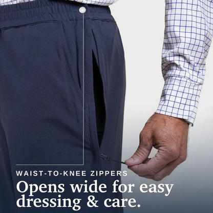 Close-up of a person wearing dark navy pants with a hand demonstrating the wide opening of the waist-to-knee zipper. A white and blue checkered shirt sleeve is visible, and text reads 'WAIST-TO-KNEE ZIPPERS' and 'Opens wide for easy dressing & care. Joe & Bella CareZips® Men's Access Pants #Dark Navy