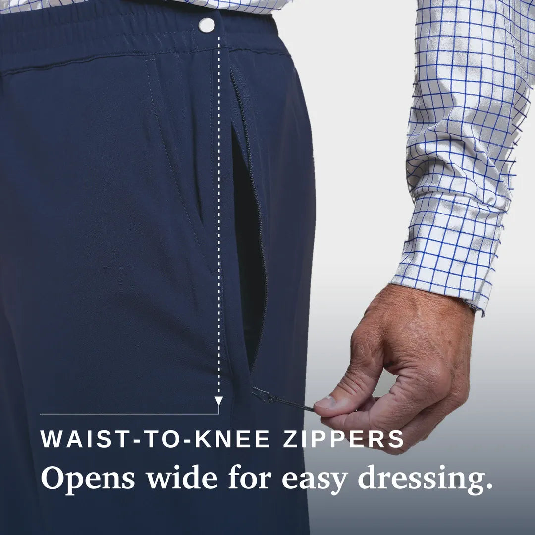 Close-up of a person wearing dark navy pants with a hand demonstrating the wide opening of the waist-to-knee zipper. A white and blue checkered shirt sleeve is visible, and text reads 'WAIST-TO-KNEE ZIPPERS' and 'Opens wide for easy dressing & care. Joe & Bella CareZips® Men's Access Pants #Dark Navy