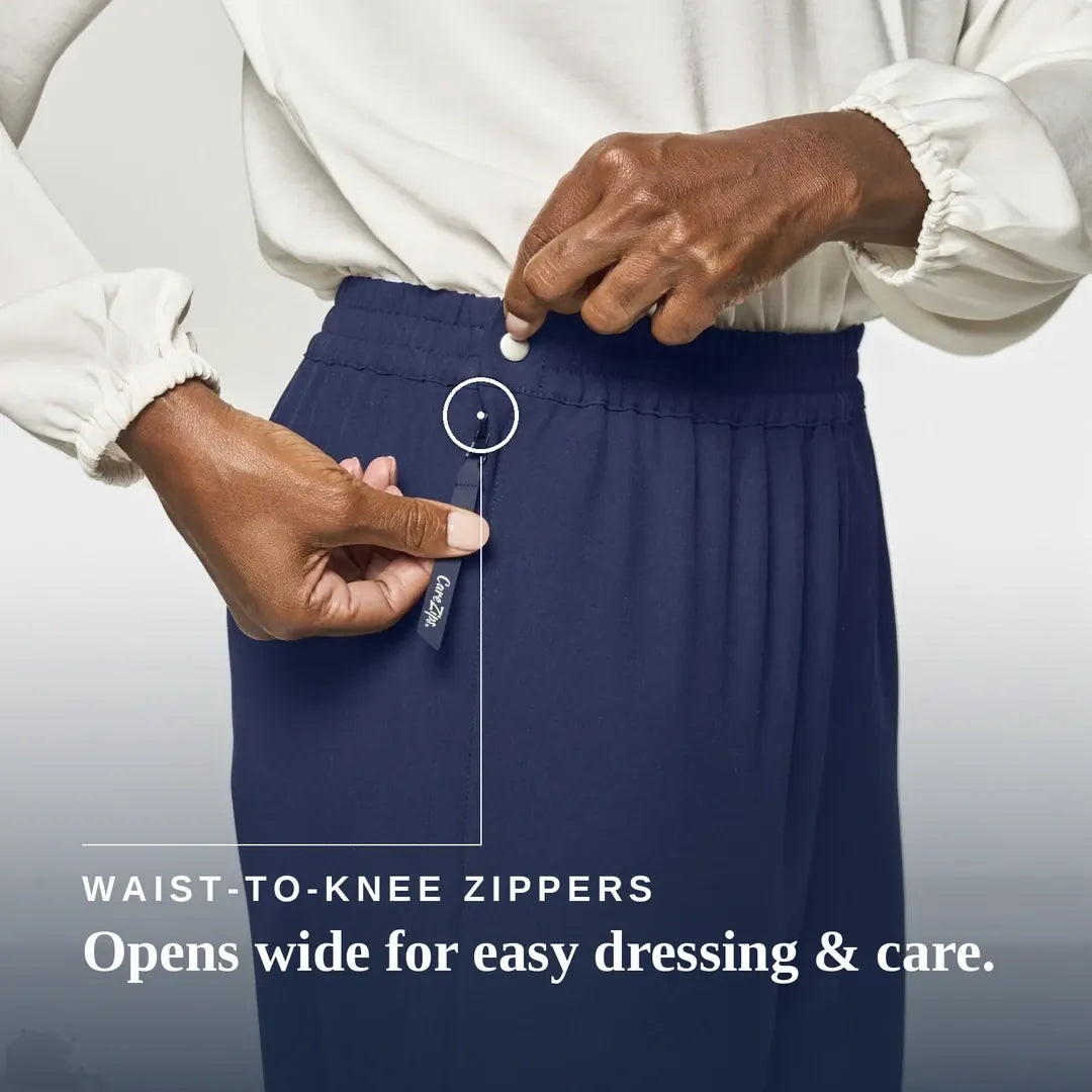 A close-up of a person wearing navy blue waist-to-knee zippers pants, demonstrating how easily the waistband can be opened. The person is wearing a white long-sleeve blouse, and the image includes the text 'WAIST-TO-KNEE ZIPPERS Opens wide for easy dressing & care. Joe & Bella CareZips® Women's Access Pants #Navy