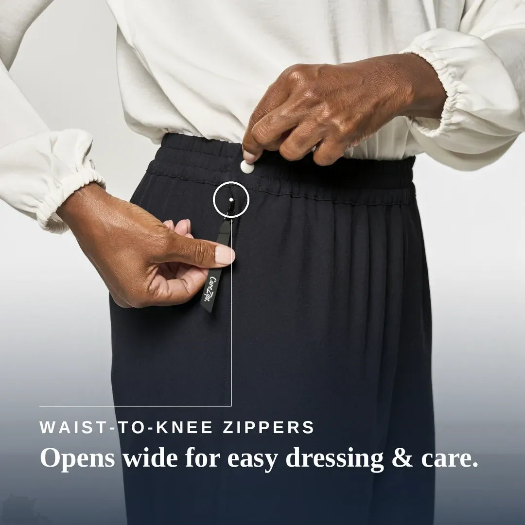 A close-up of a person wearing dark navy pleated trousers and a white blouse, demonstrating a waist-to-knee zipper. The person's hands are shown opening the zipper, with a small black fabric tag attached to the zipper pull. Joe & Bella CareZips® Women's Access Pants #Black