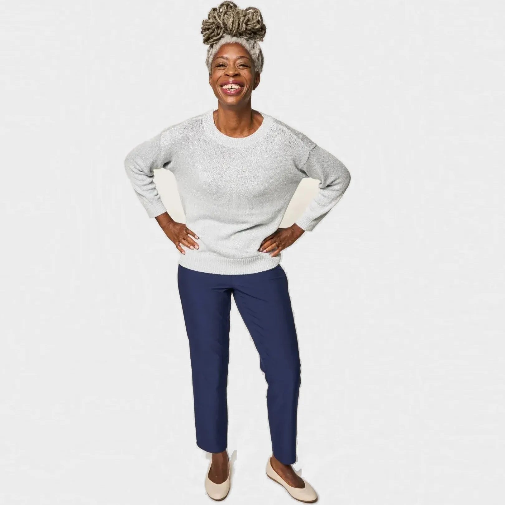 ["A smiling woman stands against a light gray studio background wearing a light gray long-sleeve knit top, navy blue pants, and beige flats. She has a patterned hair bun and is posing with her hands on her hips. Joe & Bella CareZips® Women's Adaptive Pants ", "Navy"]