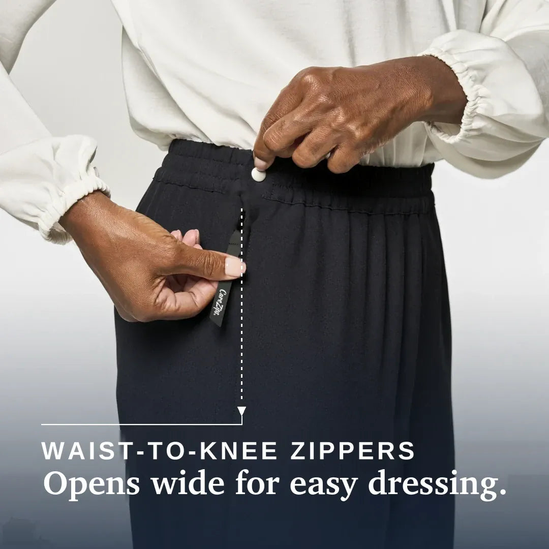 ["A close-up of a person wearing dark navy pleated trousers and a white blouse, demonstrating a waist-to-knee zipper. The person's hands are shown opening the zipper, with a small black fabric tag attached to the zipper pull. Joe & Bella CareZips® Women's Access Pants ", "Black"]