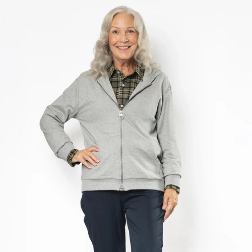 An older woman stands against a plain white studio background wearing a light gray zip-up hoodie over a green plaid shirt, dark navy pants, and cream-colored sneakers. #Grey