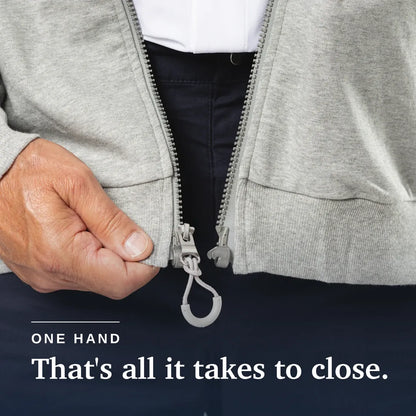Close-up of a person's hand closing a light gray zip-up jacket with a built-in carabiner, over dark trousers and a white shirt. Text at the bottom reads 'ONE HAND That's all it takes to close. Joe & Bella HoodEase™: The Magnetic-Zip-Up Hoodie #Grey