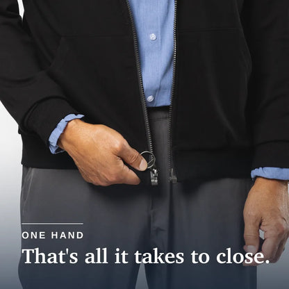 A close-up of a person wearing a black zip-front jacket over a light blue dress shirt and gray trousers, with one hand pulling the zipper tab to close the jacket. White text at the bottom reads 'ONE HAND That's all it takes to close. Joe & Bella HoodEase™: The Magnetic-Zip-Up Hoodie  #Black