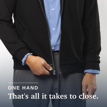 A close-up of a person wearing a black zip-front jacket over a light blue dress shirt and gray trousers, with one hand pulling the zipper tab to close the jacket. White text at the bottom reads 'ONE HAND That's all it takes to close. Joe & Bella HoodEase™: The Magnetic-Zip-Up Hoodie  #Black