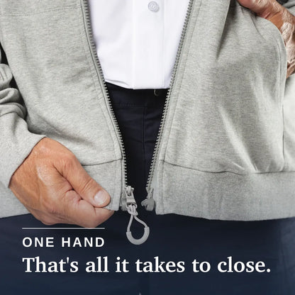 Close-up of a person's hand closing a light gray zip-up jacket with a built-in carabiner, over dark trousers and a white shirt. Text at the bottom reads 'ONE HAND That's all it takes to close. Joe & Bella HoodEase™: The Magnetic-Zip-Up Hoodie #Grey