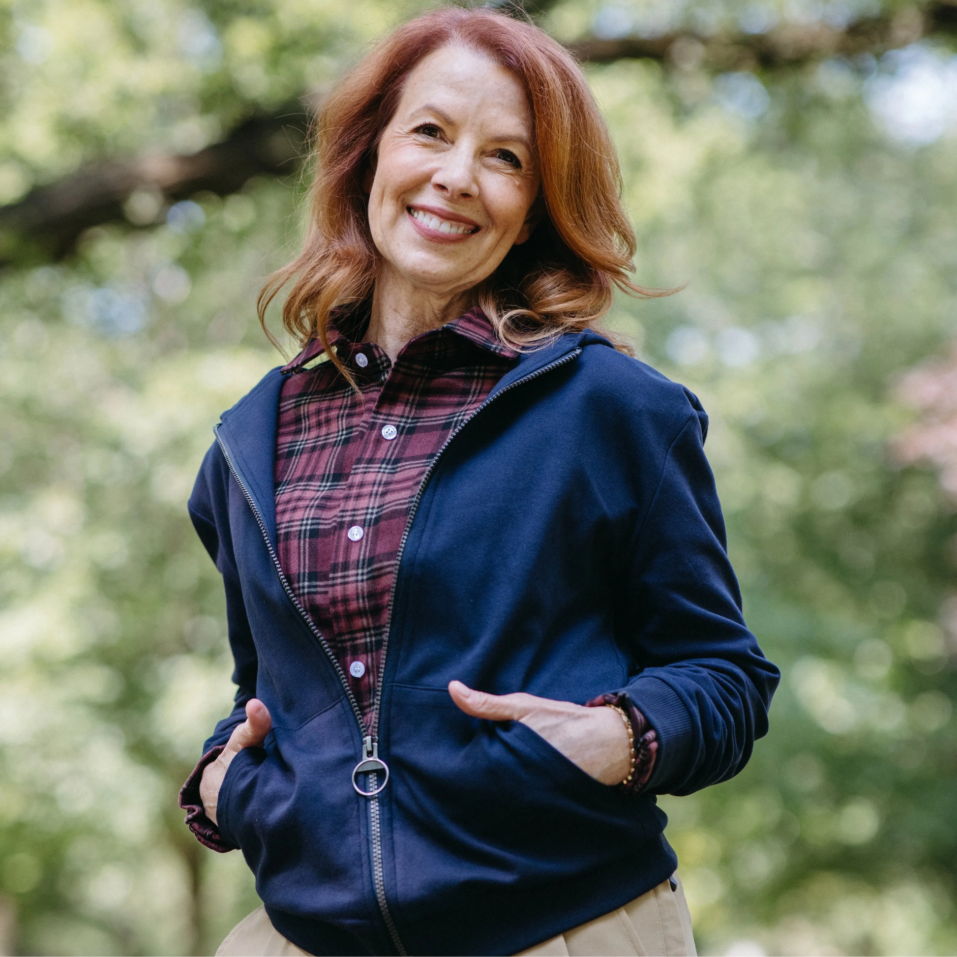 ["A smiling woman with red hair stands outdoors in a park wearing a navy zip-up hoodie over a red and black plaid button-down shirt, paired with beige pants. Navy blue hoodie with a zipper on a beige background ", "Navy"]