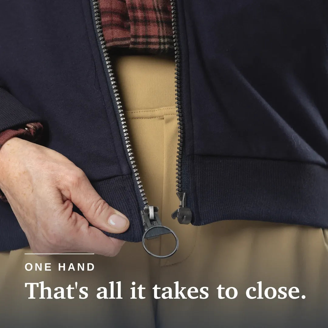 Close-up of a person's hand closing a dark navy zippered jacket, with tan pants and a red plaid shirt visible underneath. White text at the bottom reads 'ONE HAND That's all it takes to close. Navy blue hoodie with a zipper on a beige background #Navy