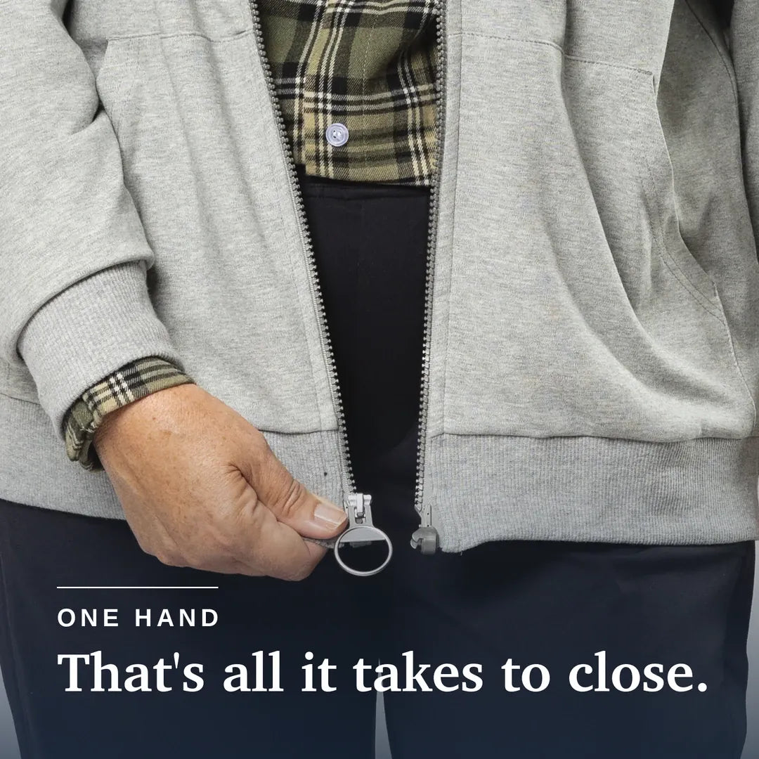 Close-up of a person wearing a light gray zip-up hoodie and dark pants, using one hand to pull the zipper closed. A plaid shirt is visible underneath, and white text at the bottom reads 'ONE HAND That's all it takes to close. Joe & Bella Heathered Grey / S HoodEase™: The Magnetic-Zip-Up Hoodie for Women #Grey