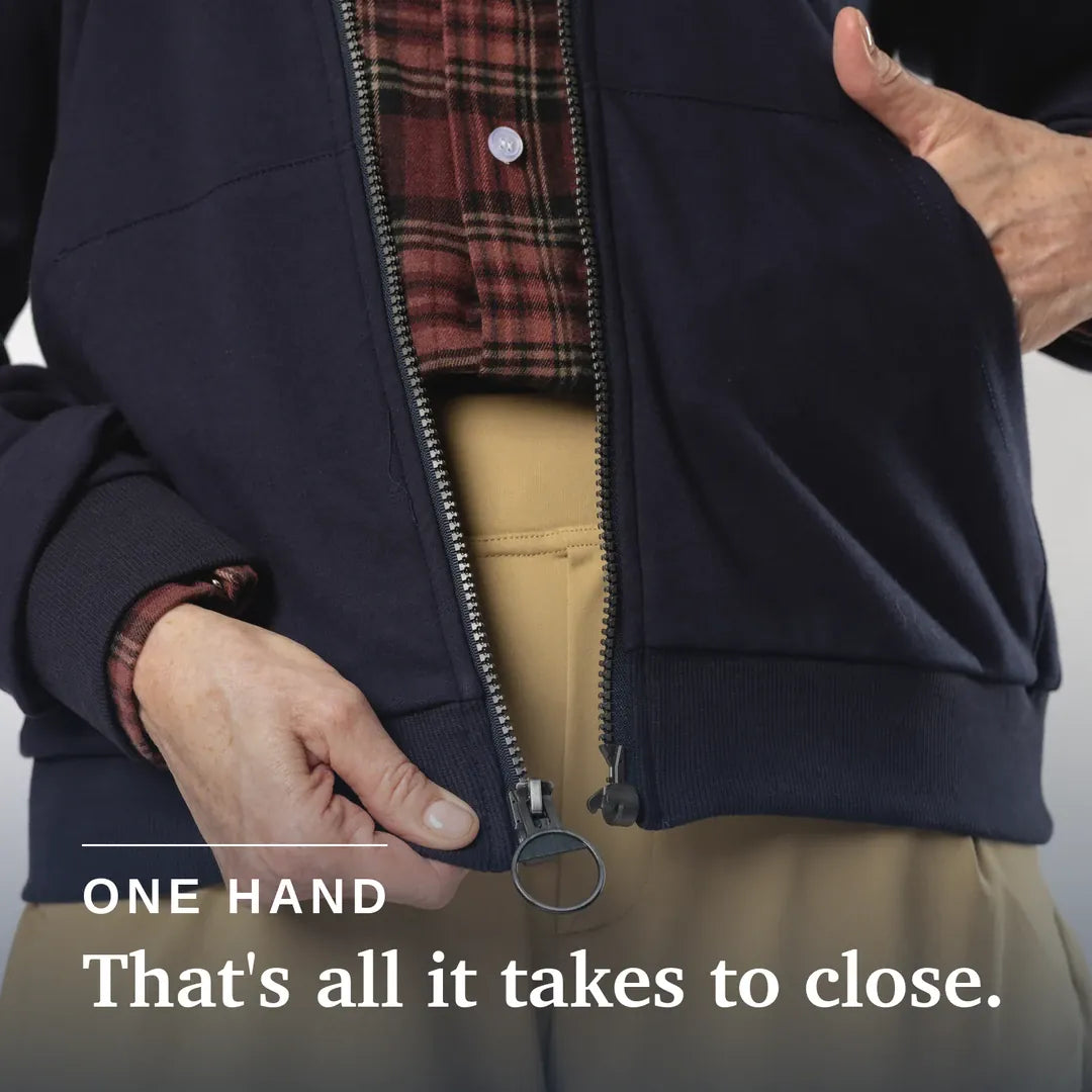 Close-up of a person's hand closing a dark navy zippered jacket, with tan pants and a red plaid shirt visible underneath. White text at the bottom reads 'ONE HAND That's all it takes to close. Navy blue hoodie with a zipper on a beige background #Navy