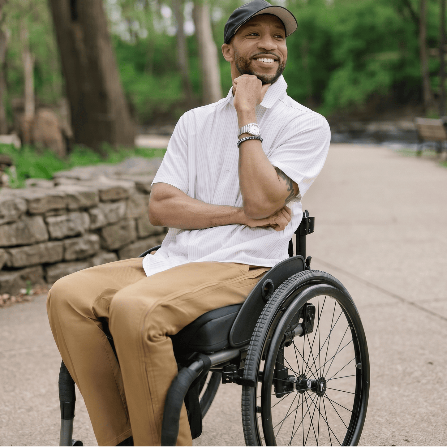 Man in a wheelchair sitting on a path in a park with trees and a bench in the background. #Khaki