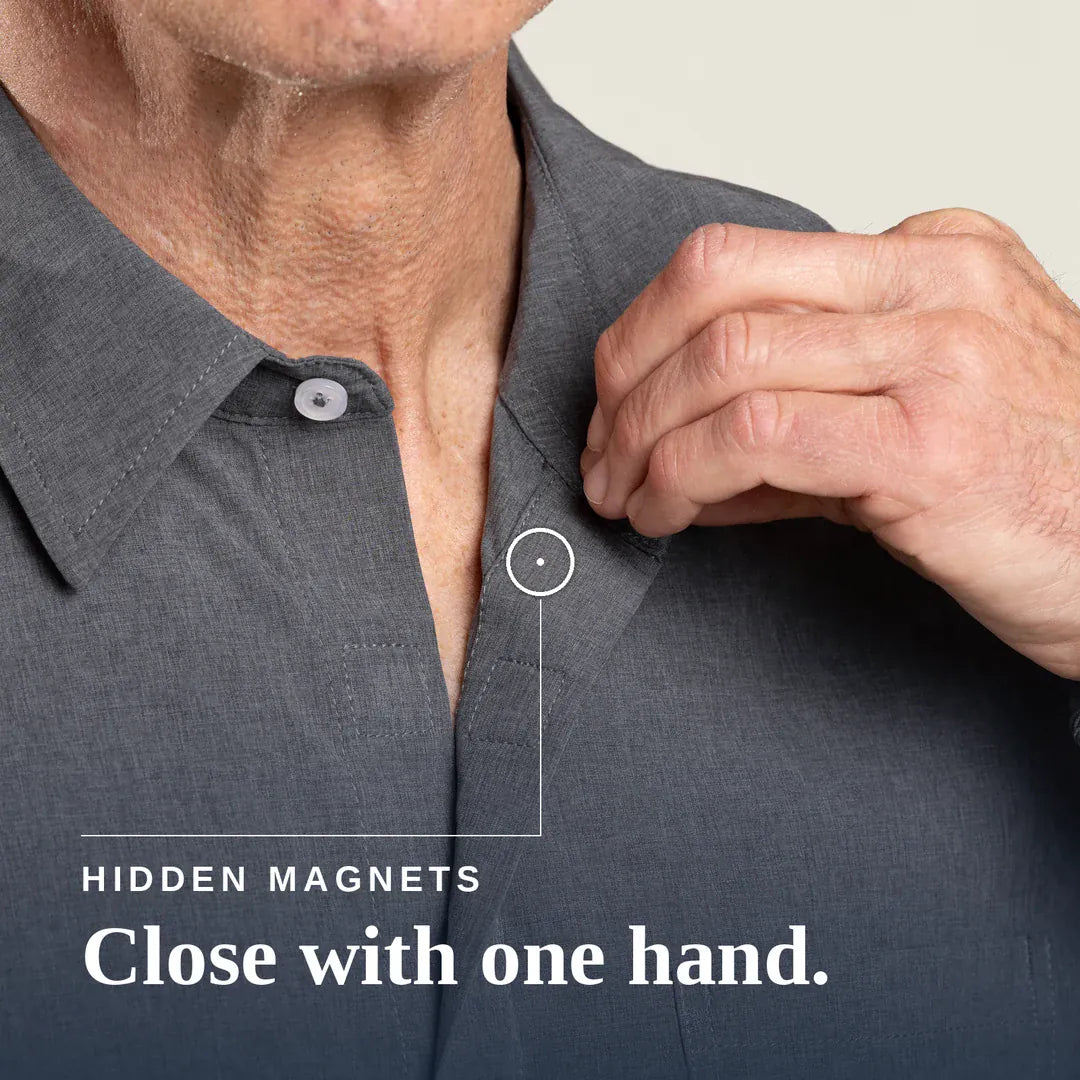 Close-up of a man's hand pulling open a dark gray button-down shirt near the collar, with text reading 'HIDDEN MAGNETS Close with one hand.'. Joe & Bella Everyday Magnetic Button-Down for Men | Relaxed-Fit #Heathered Grey