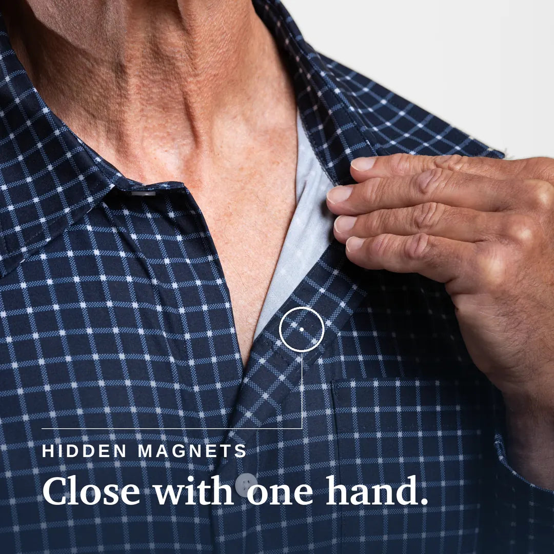 Close-up of a man's neck and chest wearing a navy and white plaid button-up shirt, with his hand reaching toward a small hidden magnet near the collar. Text on the image reads 'HIDDEN MAGNETS Close with one hand. Joe & Bella Everyday Magnetic Button-Down for Men | Relaxed-Fit #Navy Check
