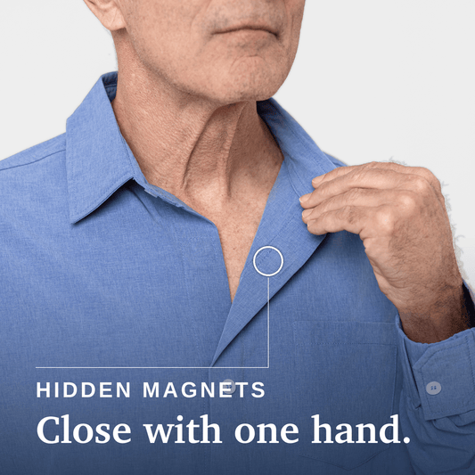 A man wearing a light blue long-sleeve button-up shirt over a white t-shirt, standing in front of a modern glass door and brick wall. #Heathered Blue