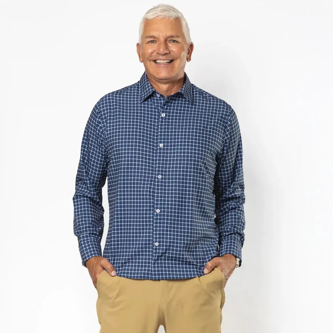 A smiling older man standing against a clean white studio background, wearing a blue and white checkered button-down shirt, tan chinos, and white sneakers. #Navy Check