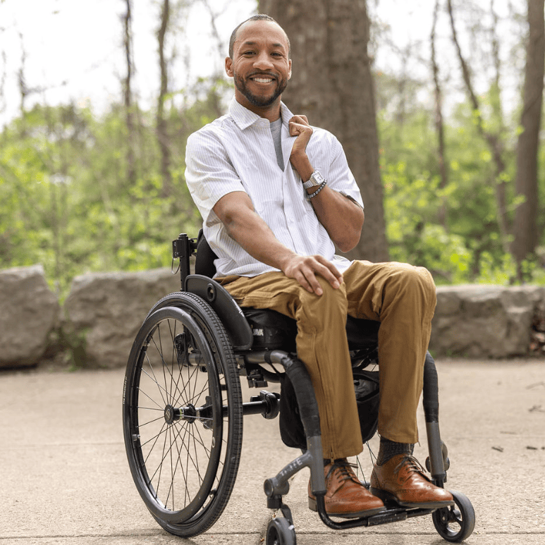Man in a wheelchair sitting outdoors with trees and rocks in the background #Tan Stripe