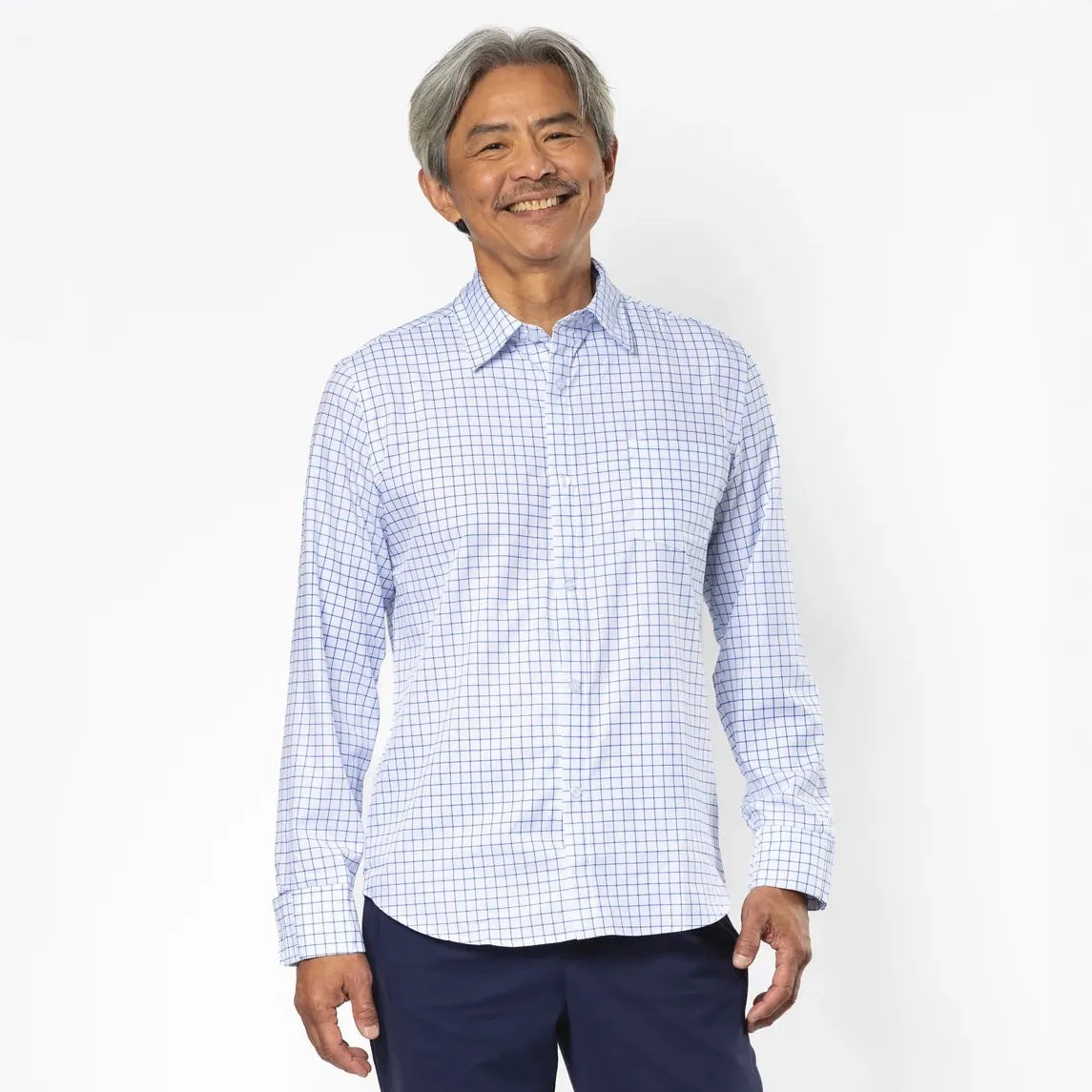 An older man stands against a plain white studio background wearing a light blue and white gingham long-sleeve button-down shirt, navy blue trousers, and beige sneakers. #White Check